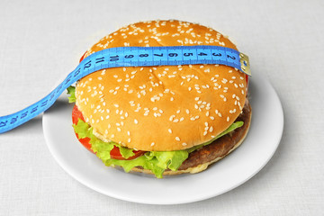Dieting concept. Plate with tasty hamburger and measuring tape on light background