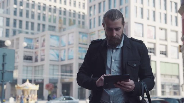 4K, Young man with beard and ponytail using tablet while standing on street.