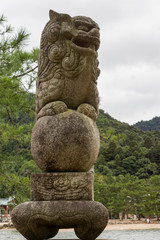 Hiroshima, Japan - September 20, 2016: Historic old brown stone statue of lion-like monster outside itsukushima Shinto Shrine. Acts as guardian. Beast sits on sphere. Green foliage background.