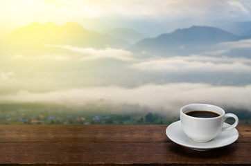 A Cup of coffee  on a wooden table with mountain, fog and sunflare blurred background in morning.