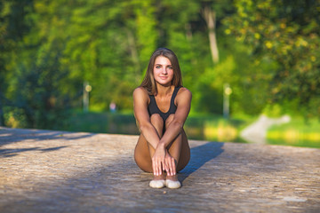 Cute girl makes sports exercises on the nature background. She wore a black kombidress body.She sits facing us. Girl smiling