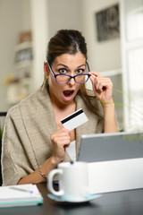 Surprised woman shopping online on laptop at home. Amazed female with glasses opening mouth and holding credit card.