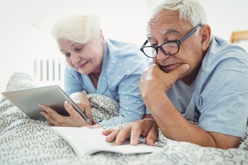 Senior couple reading a book and using digital tablet on bed