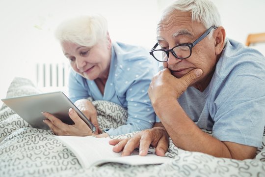Senior couple reading a book and using digital tablet on bed