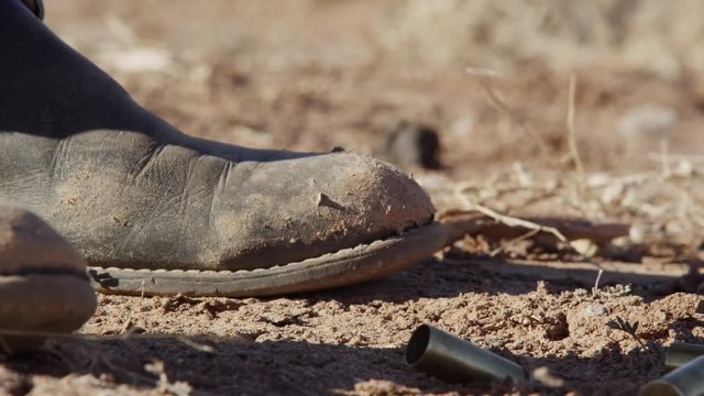 Closeup of a mans boot with bullet shell casings falling on the dirt
