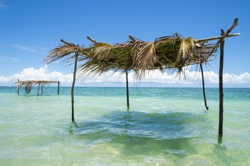 Fototapeta na wymiar Rustic palm frond and tree branch palapa umbrella stands waiting to shade visitors to the shallow waters on a remote tropical beach in Nordeste northeastern Bahia, Brazil