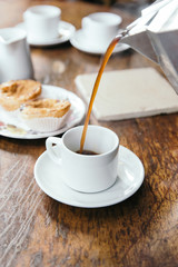 Black coffee pouring into white cup with cakes on wooden table.
