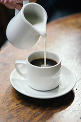 Milk pouring into black coffee on wooden table.