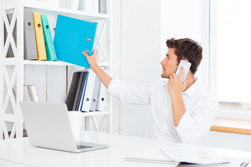 Young businessman taking folder from shelf and talking on phone