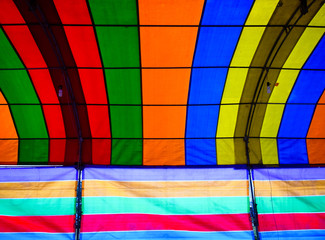Inside a colorful tent. The tent consists of green, red, orange, blue, yellow and purple colors and lines form repetitive patters of color strips. There are lamps hanging down the tent props.