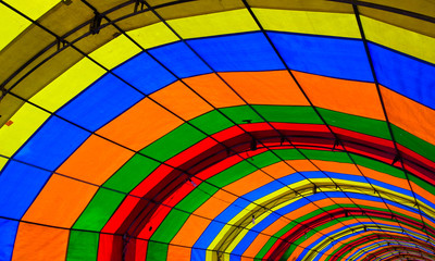 A  photo taken inside a colorful tent. The tent consists of green, red, orange, blue and yellow colors and form repetitive patters of color strips. There are lamps hanging down the tent props.