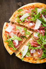 Sliced pizza with prosciutto (parma ham), arugula (salad rocket) and parmesan on dark wooden...