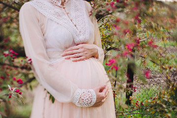 Intentionally blurred image of a pregnant caucasian woman in beautiful light pink dress holding her hands on her belly in the park. Tilt-shift lens used, soft selective focus.