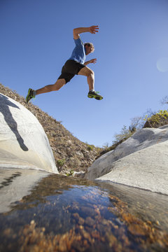 An Adult Man Jumping From One Rock To Another Over The Top Of The Waterfall At Sierra De La Laguna.  Baja California, Mexico.