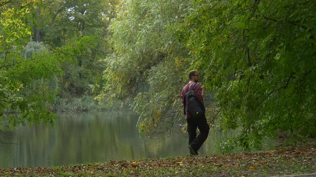 Backpacker Comes Sits Down at the Lake Bank Park Forest Walking and Looking at Smooth Water Resting Outdoors Dry Leaves on a Ground Autumn Landscape
