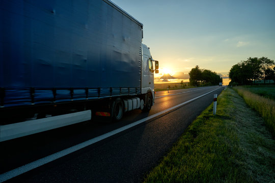 Truck driving on asphalt road towards the horizon in rural landscape at sunset.