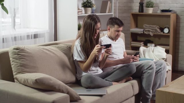 Young couple sitting on sofa in apartment looking at mobile phones and not talking to each other