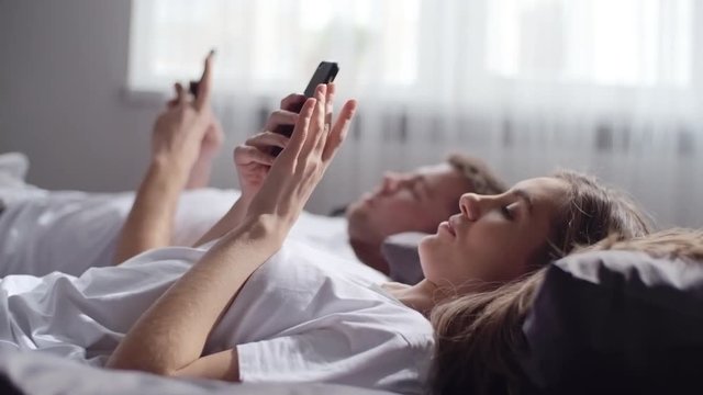 Handheld shot with rack focus of young man and woman lying in bed in morning and looking at their mobile phones