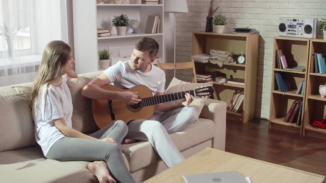 Young laughing woman with long hair sitting on sofa beside her boyfriend and listening to him playing guitar to her and singing