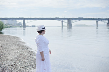 A woman in a white dress and a hat standing on the beach and looking into the distance.