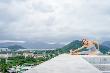 Fototapeta na wymiar Yoga on rooftop. Happy young woman stretching on roof with city and mountains view.