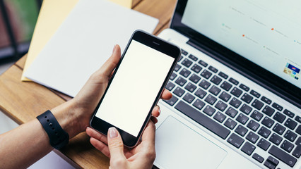 Close-up of smartphone with blank screen in female hands. Girl presses finger on smartphone button. In background, the laptop on a wooden table, a letter and an envelope. She uses a digital gadget.