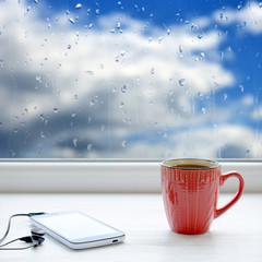 Cup of coffee, smartphone and headphones on a windowsill. In the background window with raindrops and clouds