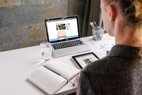 Back view. Business woman in gray shirt sits at white desk.On table is 
laptop,on-screen page online store,near notebook,digital tablet,glass of
 water and pencil holders.Woman working in office.