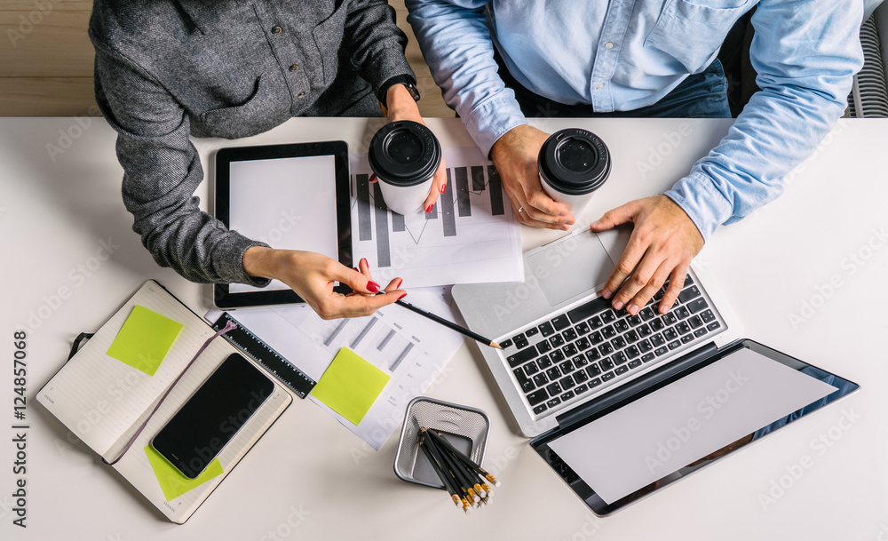 Wall mural View from above. Businessman holding cup of coffee and printing. Businesswoman showing pencil on screen of laptop. Teamwork. On table smartphone,digital tablet, graphics, notebook, pencil holders.