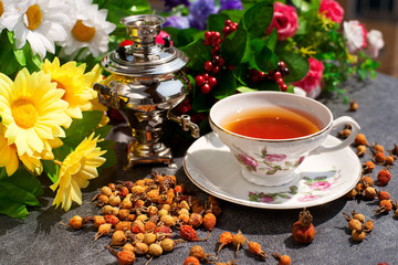 Лечебный чай с ягодами шиповника на фоне цветов. Чай в самоваре создает домашний уют. Солнечные лучи красиво освещают чай и ягоды шиповника.