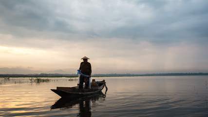 Fototapeta na wymiar Asian fisherman on wooden boat casting a net for catching freshwater fish in nature river in the early morning before sunrise