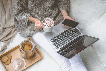 Woman in knit sweater sitting in cozy atmosphere of home and holding smartphone.Notebook is on girl's lap.Nearby stands tray with cup of cocoa with marshmallows and jar of cookies.Girl using gadget.
