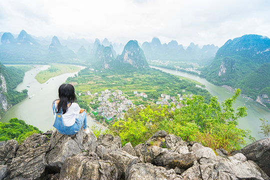 Young woman sitting on the top of mountain and enjoing view on valley with rocks. Xing Ping, Yangshuo, China.