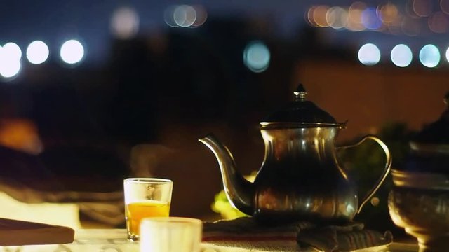 Traditional Metal Pot and Glasses With Steaming Moroccan Tea on the Night City View Rooftop Terrace, Midshot, Night Light