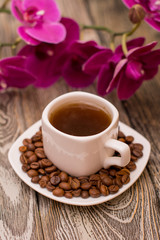 Small white cup of coffee, roasted coffee beans, orchid on wooden background