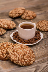 Small white cup of coffee, roasted coffee beans, cookies with sesame seeds on wooden background