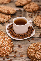 Small white cup of coffee, roasted coffee beans, cookies with sesame seeds on wooden background