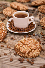 Small white cup of coffee, roasted coffee beans, cookies with sesame seeds on wooden background