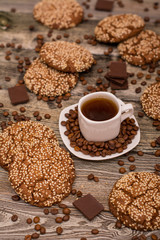 Small white cup of coffee, roasted coffee beans, cookies with sesame seeds, slices of chocolate on wooden background