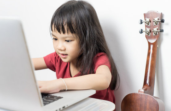 Little Musician Kid Is Working On Her Music On A Laptop. Asian Kid Is Looking For Music Note On The Internet. Japanese Girl Is Taking A Guitar Music Course Online.