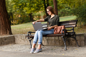 Young female student sitting on a bench in park, she is working on laptop.