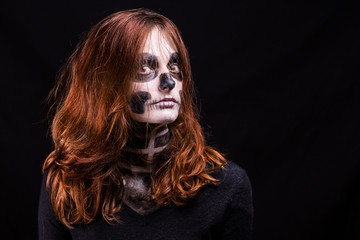 Une femme à la chevelure rousse vif et maquillée en squelette pose sur fond noir en nous regardant