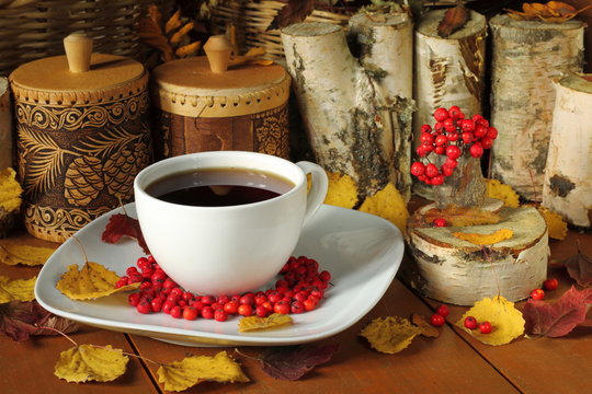 На деревянном столе расположены кружка с чаем, ягоды рябины, осенние опавшие листья и домашняя утварь.
