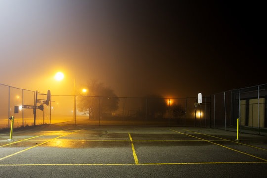 A fenced tennis and basketball court at school yard on a foggy night under street lights