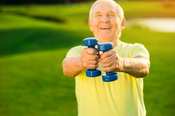 Elderly man is holding dumbbells. Old guy smiling outdoor. Effective training program. Start...