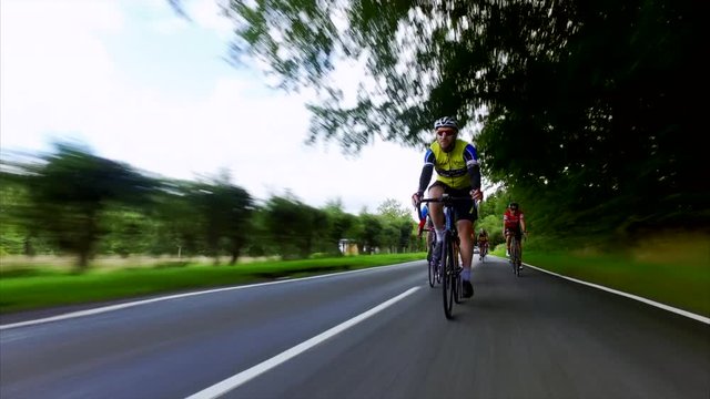 A group of cycling athletes riding fast along roads through the UK countryside on an overcast day. 