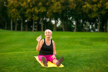 Senior lady sitting on mat. Woman smiling and holding bottle. The joy of being healthy. Take a sip.