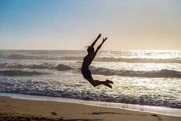 Fototapeta na wymiar Silhouette of jumping young woman on beach against crushing waves