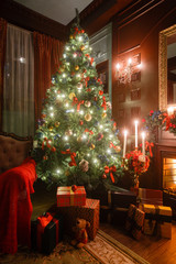 Christmas evening by candlelight. classic apartments with a white fireplace, decorated tree, sofa, large windows and chandelier.