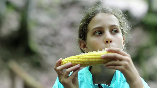 Beautiful girl in blue jacket eats corn and looks around. Girl eats yellow corn. Female with corncob
